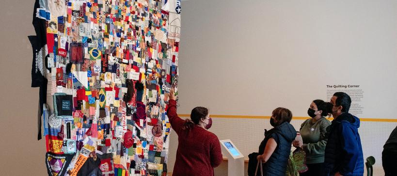 A women is pointing at a large community quilt hanging patched together with many different pieces. A family is standing next to her observing.