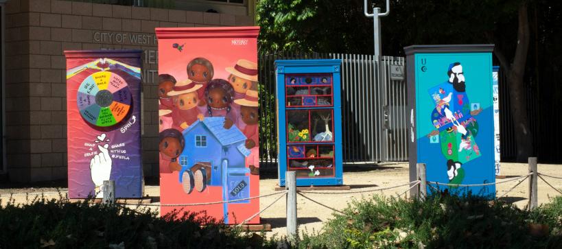 Four colorfully painted wooden boxes about 4 feet tall installed in a park.
