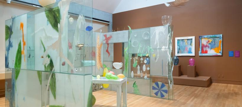 Three glass walls surround a table with colorful objects. Above the walls, larger-then-life feathers suspend from the ceiling up into a skylight.