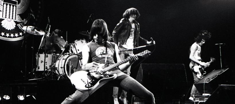 A black and white photo of a guitar player up front from his band on a stage playing.