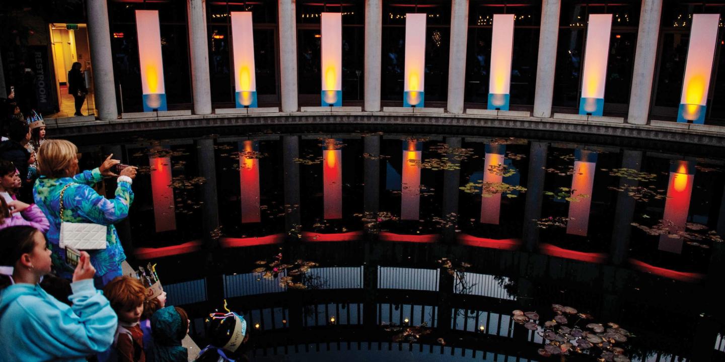 Photo of the lily pond at the Skirball lit up with large column spotlights mimicking a Hanukkah. Families stand along the pond looking at the lights.