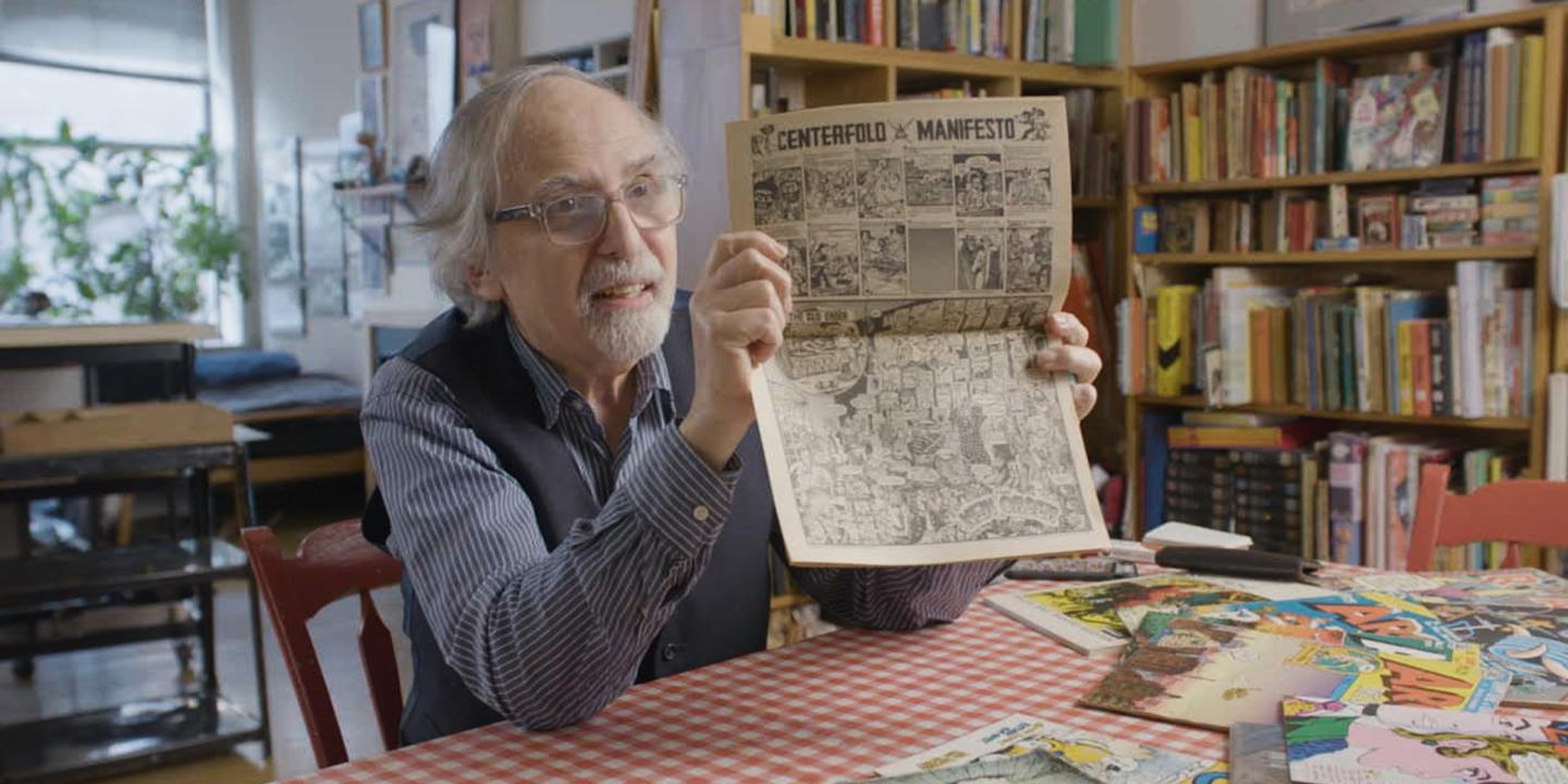 Art Spiegelman sits at a table in his home holding up a newsprint of illustration titled, Centerfold Manifesto, to the camera.