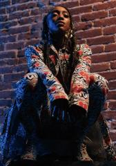 Comfort Fedoke crouches against a brick wall wearing bright, flashy clothes.