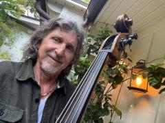 Randall Tico poses next to an upright bass in front of a white house with green plants and trees and a lantern.