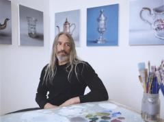 Robert Russell sits at his desk with still lifes in the background on a white wall.
