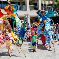 Performers on stilts in elaborate costumes move and dance in the middle of a large group of families in a Courtyard.