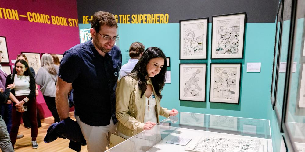A man and a woman in a gallery look at a case with comic book art in it.